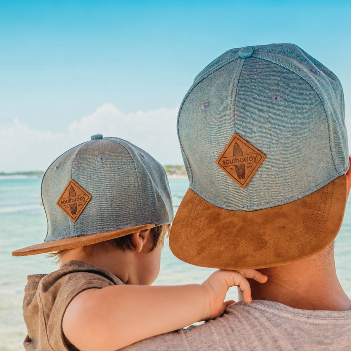 Papa und Kind von hinten mit identischen hellblauen Soulbuddy Snapback Caps am Meer