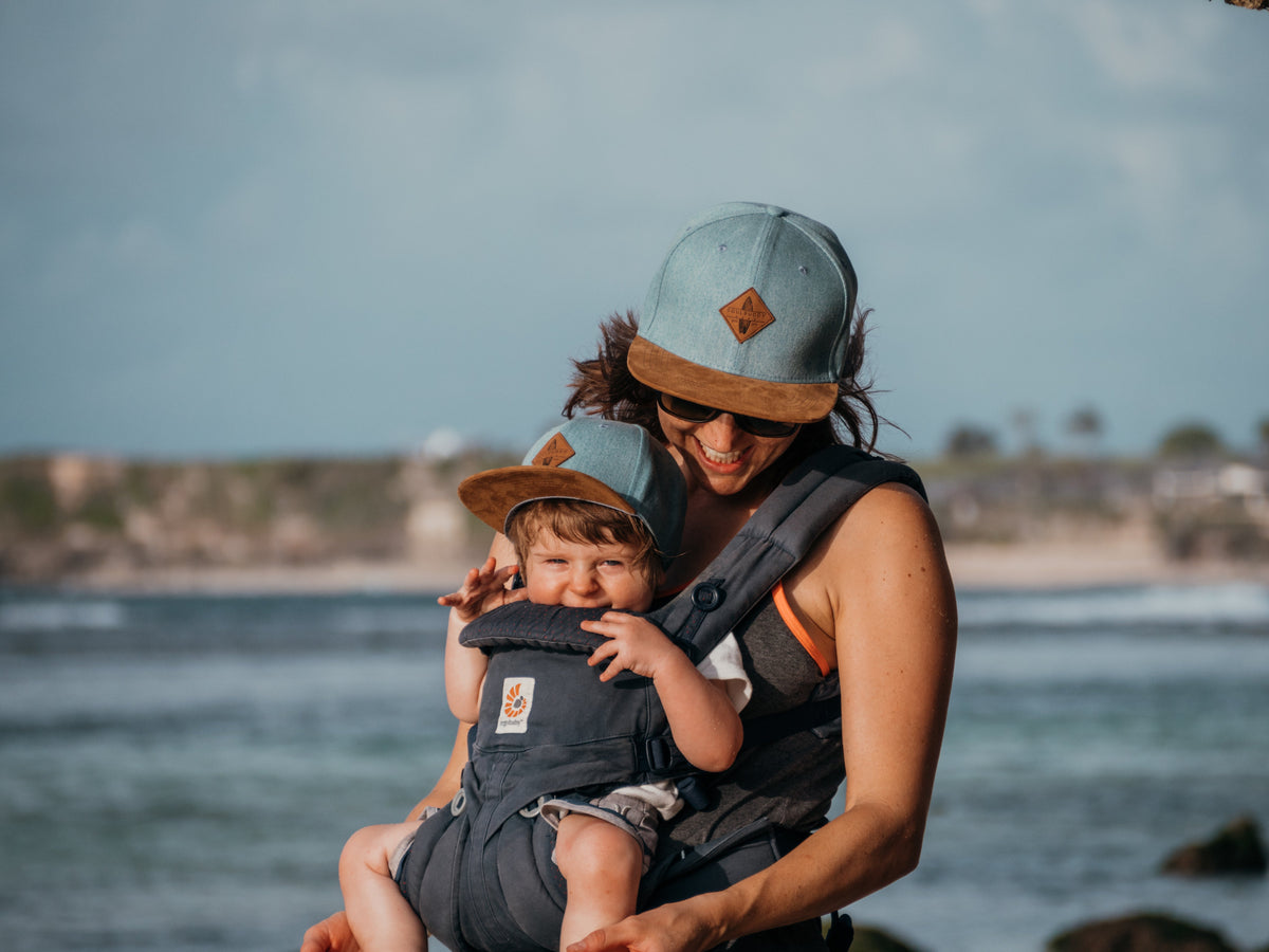 Mama mit Kleinkind in der Babytrage am Meer, beide mit Soulbuddy Snapback Caps im Partnerlook – Geschenk zum 1. Geburtstag