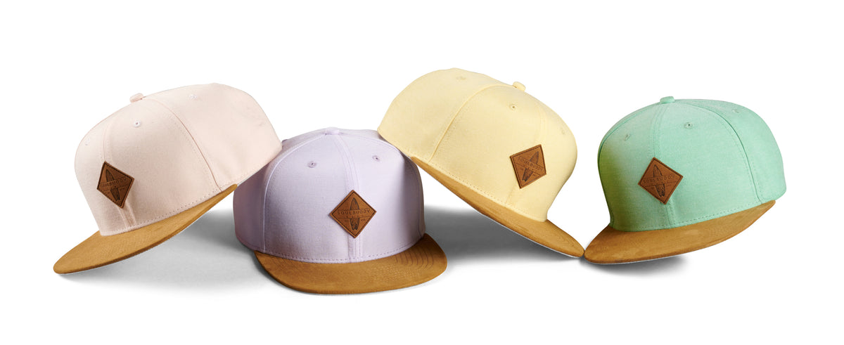 Quatre casquettes snapback Soulbuddy aux couleurs pastel côte à côte – rose, lilas, jaune et menthe avec visière en daim