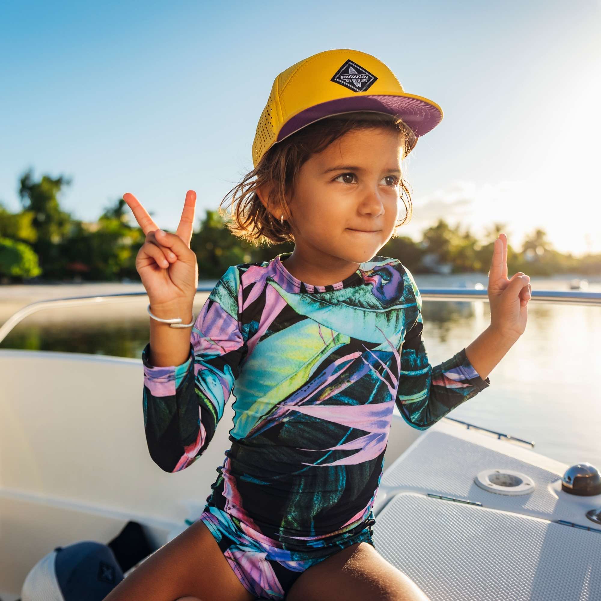 Kind mit gelber Snapback Cap zeigt fröhliche Geste auf einem Boot bei sommerlichem Sonnenuntergang am Meer
