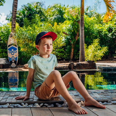Soulbuddy Kinder Performance Cap – Kind mit schwarzer Cap entspannt am Pool in tropischer Umgebung