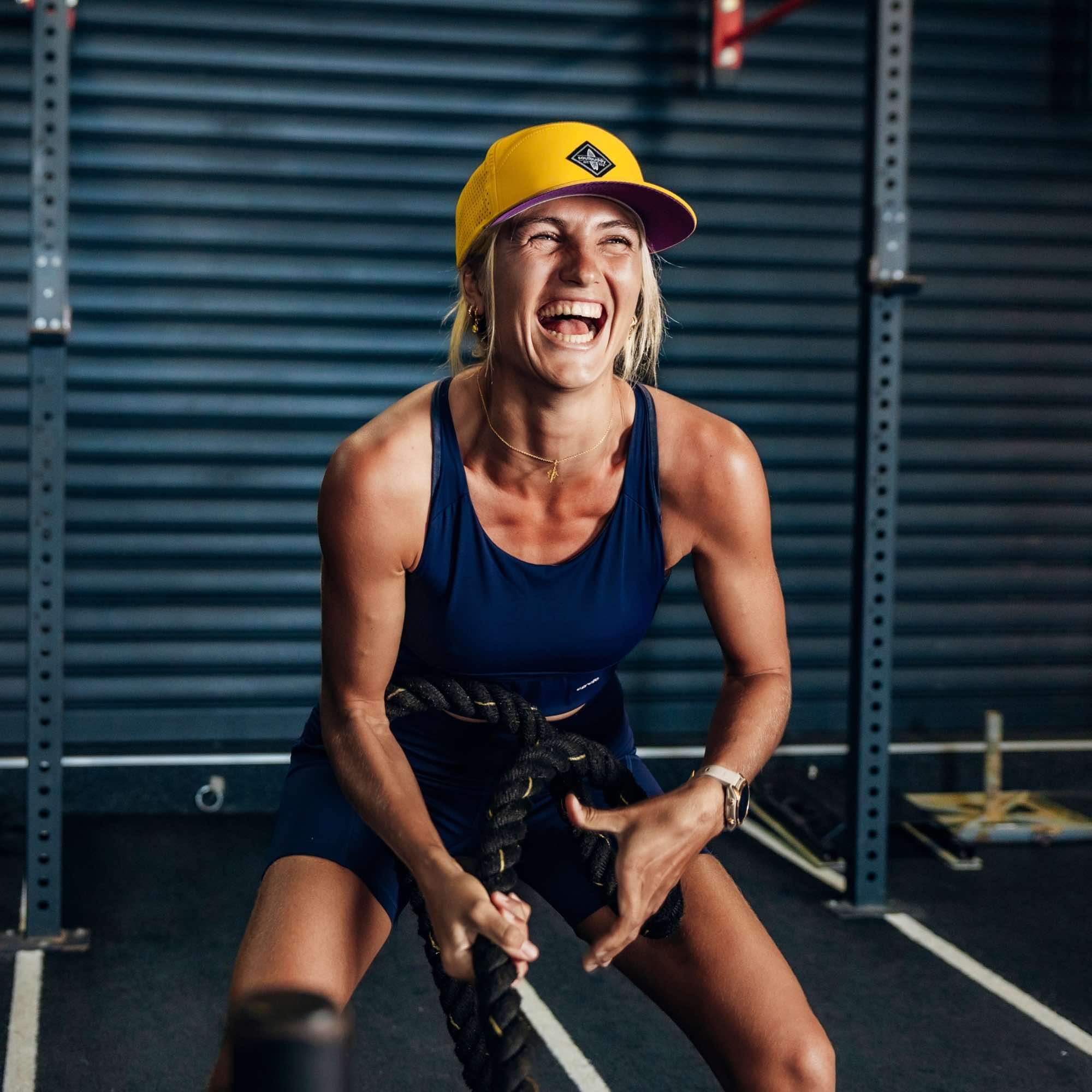 Sportliche Frau trägt gelbe Performance Snapback Cap beim intensiven Training mit Battle Ropes im Fitnessstudio