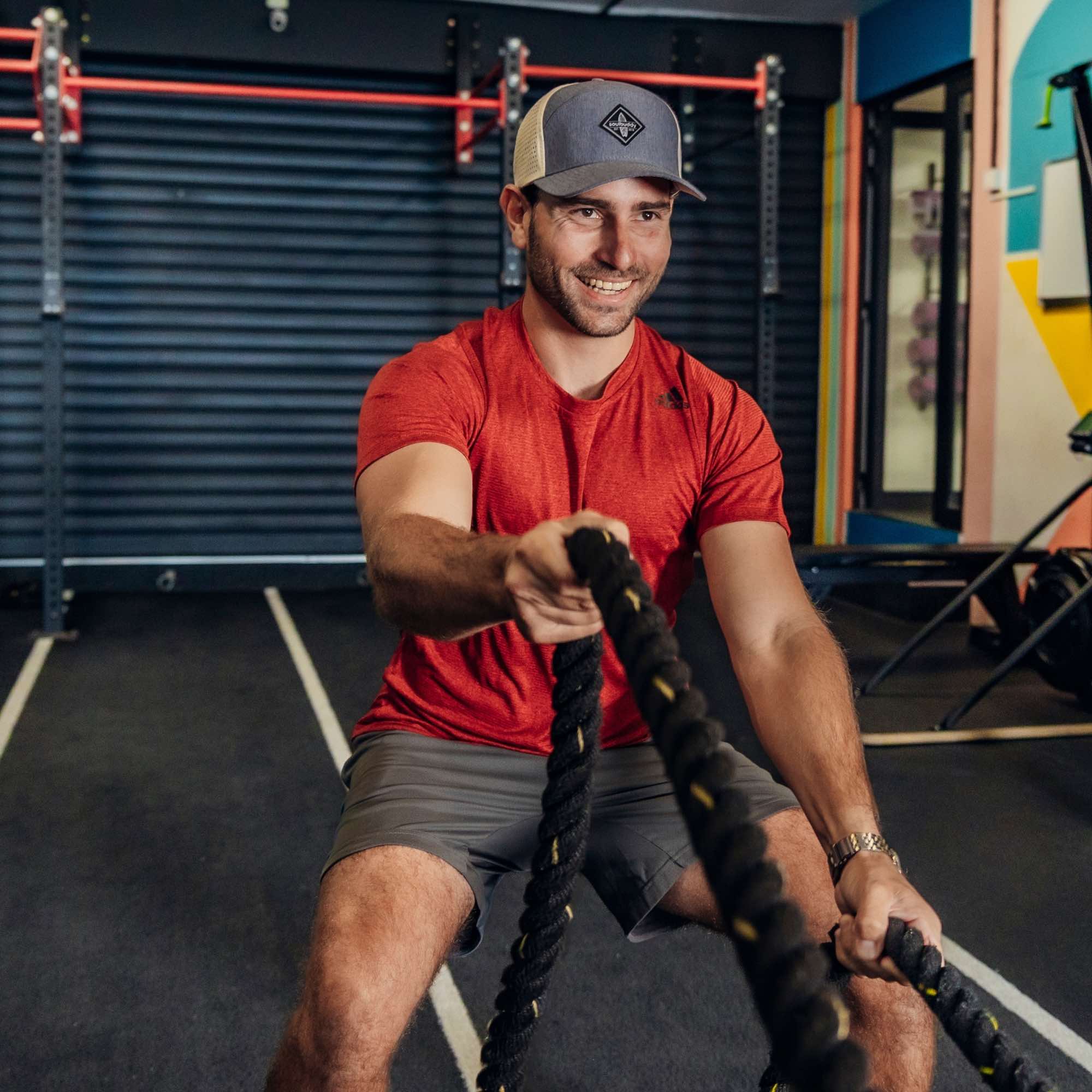 Sportlicher Mann mit grauer Snapback Cap und rotem Shirt beim Battle-Rope-Training im Fitnessstudio, trainiert mit schweren Seilen und lächelt konzentriert