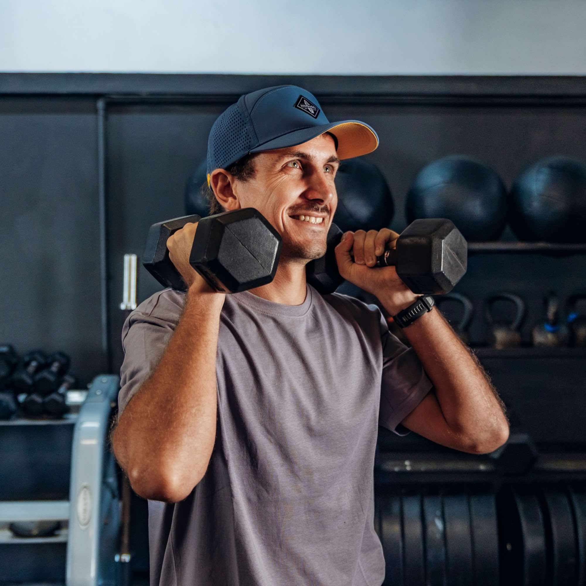 Sportlicher Mann trägt blaue Performance Snapback Cap beim Krafttraining mit Kurzhanteln im Fitnessstudio