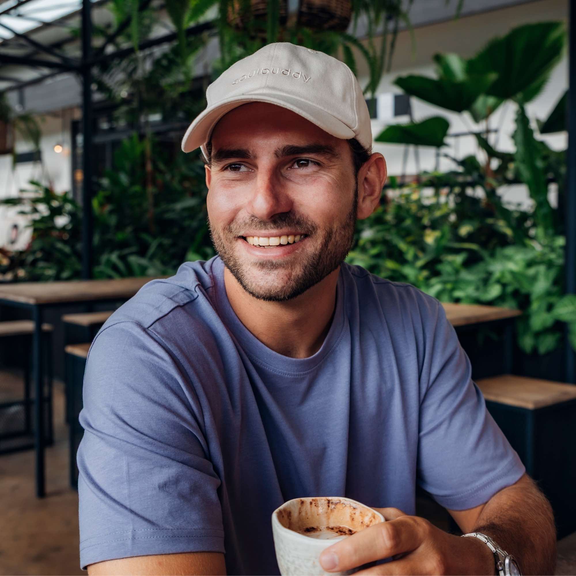 Mann mit beiger Soulbuddy Cap sitzt in einem Café und lächelt entspannt mit einer Tasse Kaffee in der Hand