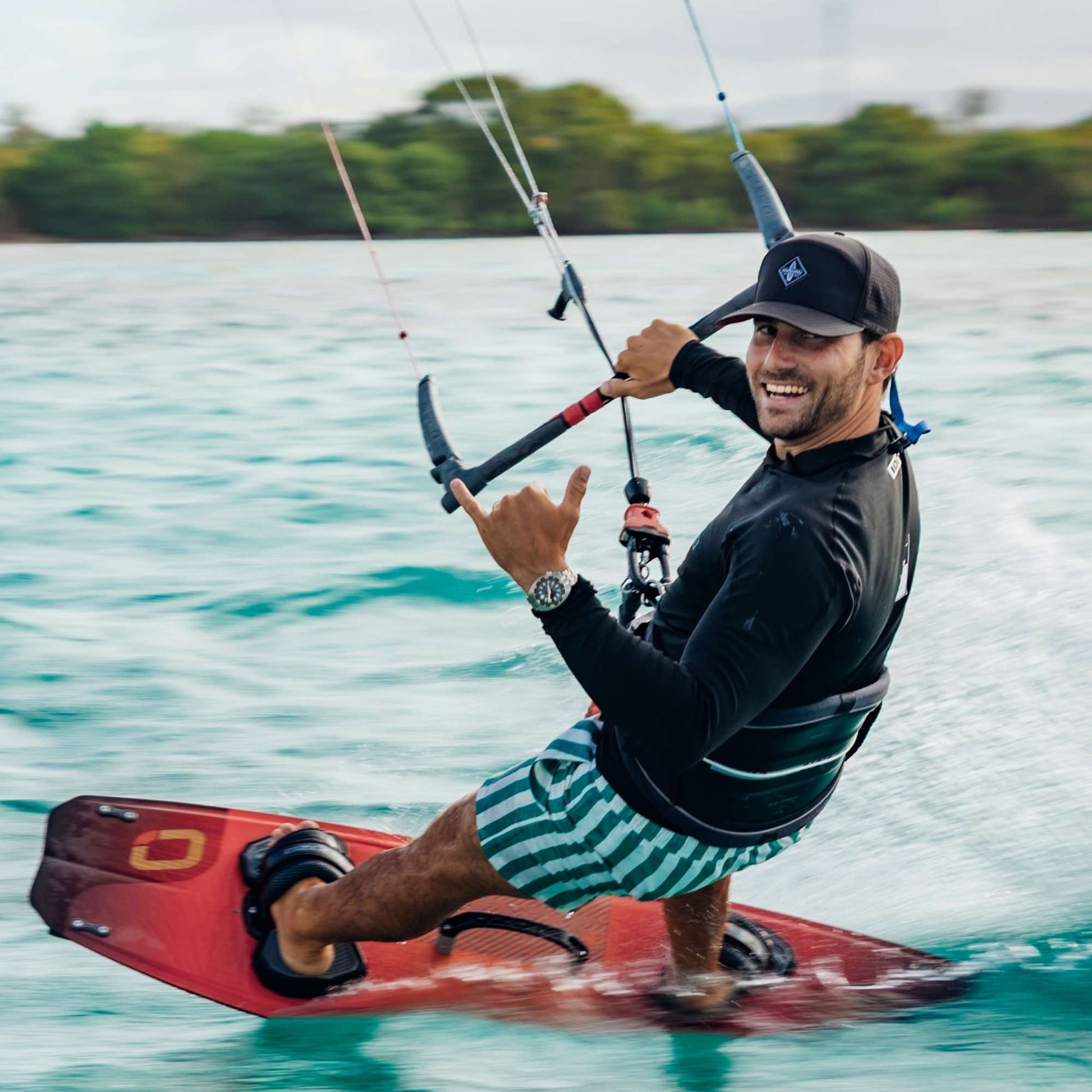Sportlicher Mann mit schwarzer Cap beim Kitesurfen auf dem Meer, hält die Bar und zeigt ein Shaka-Handzeichen, während er über das Wasser gleitet