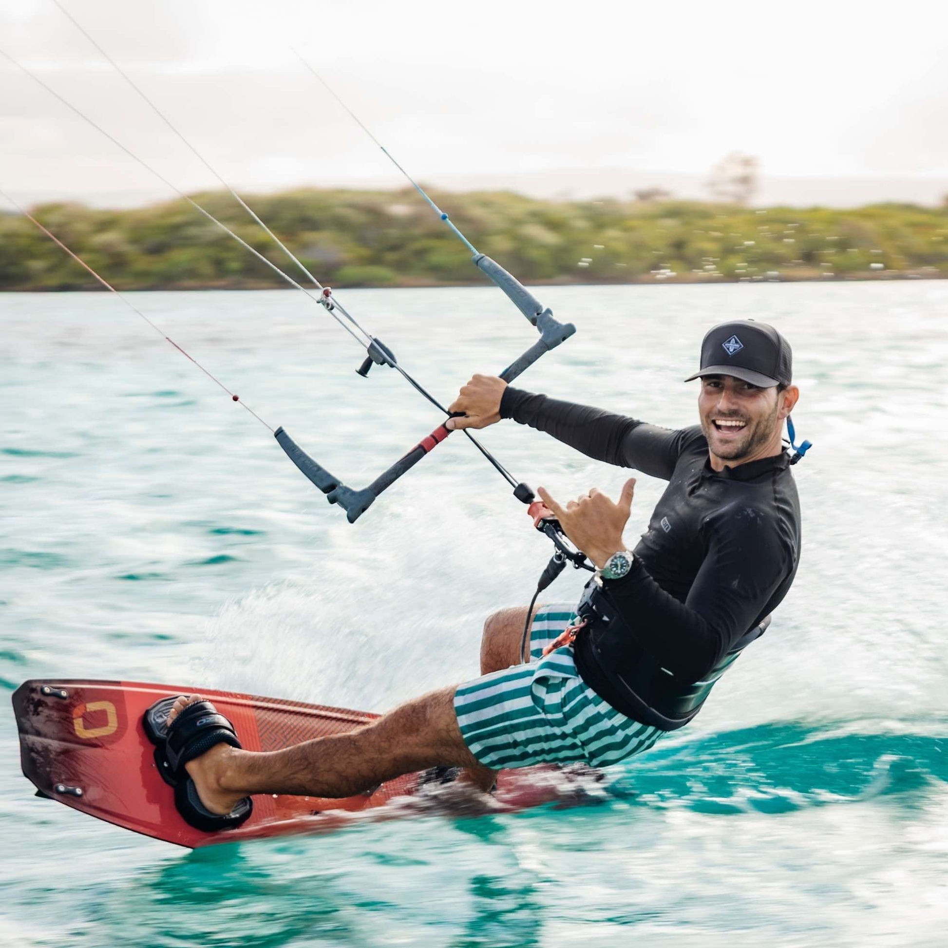 Sportlicher Mann mit schwarzer Snapback Cap beim Kitesurfen auf türkisfarbenem Wasser, macht Shaka-Handzeichen und lächelt in die Kamera