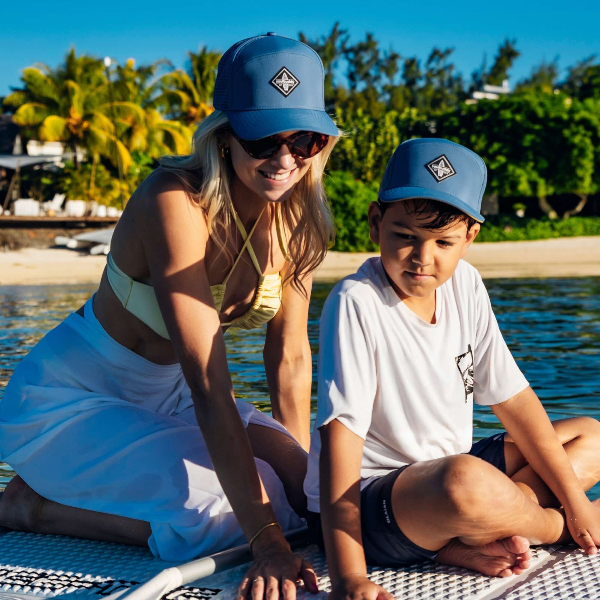 Mutter und Sohn mit blauen Snapback Caps im Partnerlook sitzen gemeinsam auf einem Stand-Up-Paddle-Board im ruhigen Wasser bei sonnigem Wetter